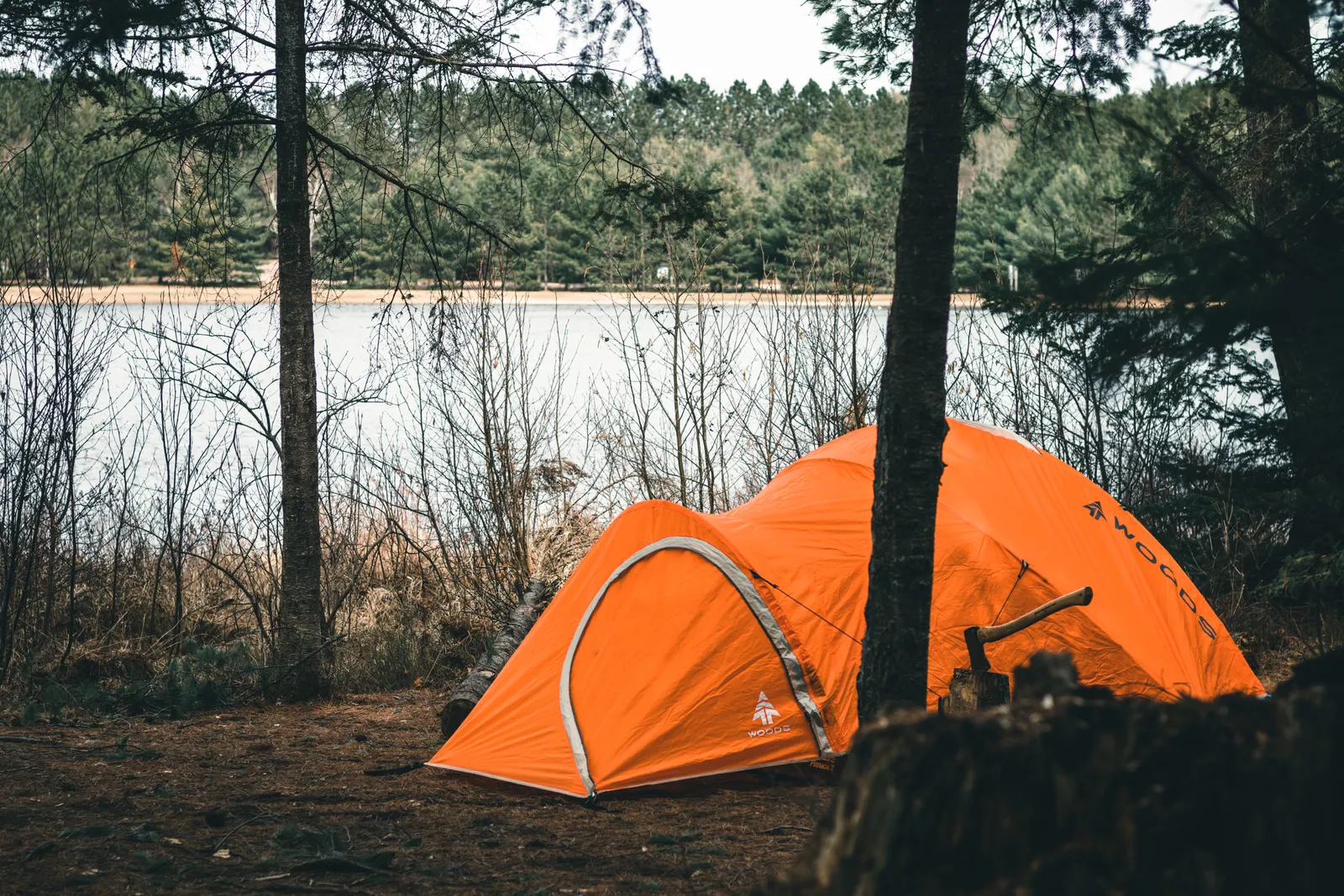 An orange tent pitched on a grassy rise beside a calm lake at the forest edge