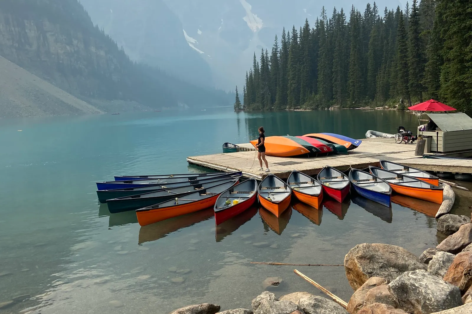 A person standing on a wooden pier beside red canoes on a glacial lake below mountains