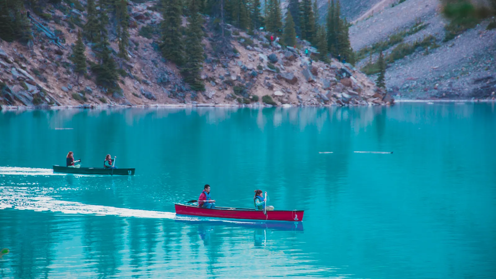 Paddlers in red canoes crossing a turquoise mountain lake surrounded by forest and peaks